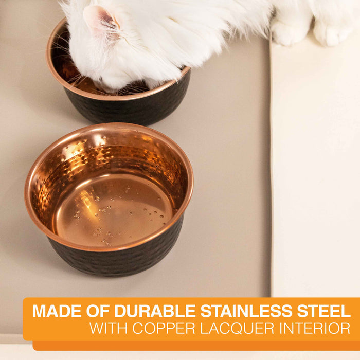 Picture of a cat eating from the Black Hammered Stainless Steel Bowl in a kitchen. Text on screen reads: Made of durable stainless steel with copper lacquer interior.
