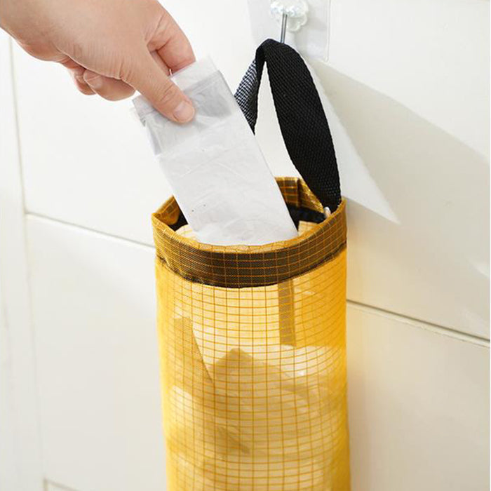 A reusable plastic bag being placed into a yellow hanging mesh bag holder mounted on a kitchen wall.